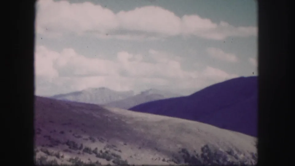 1958 Colorado 8mm frame showing open rolling hills, distant mountain ridges, sparse vegetation, and pale clouds overhead.