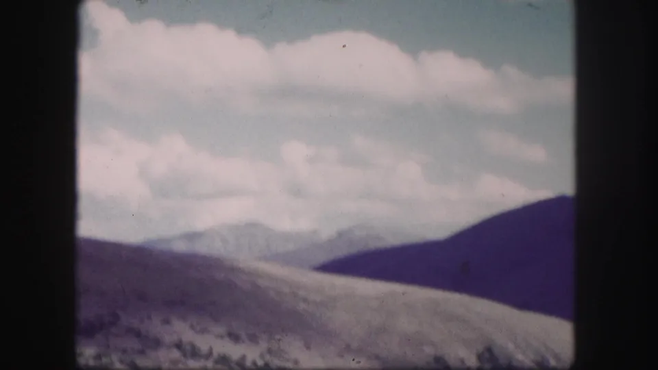 Soft 8mm Colorado view of a broad hillside, distant mountain ridge, sparse low vegetation, and white clouds in a faded sky.