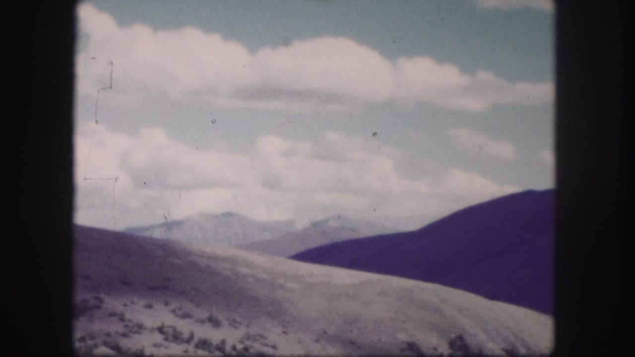 1958 Colorado 8mm frame showing distant mountain ridges, a pale hillside, and clouds across a bright sky.