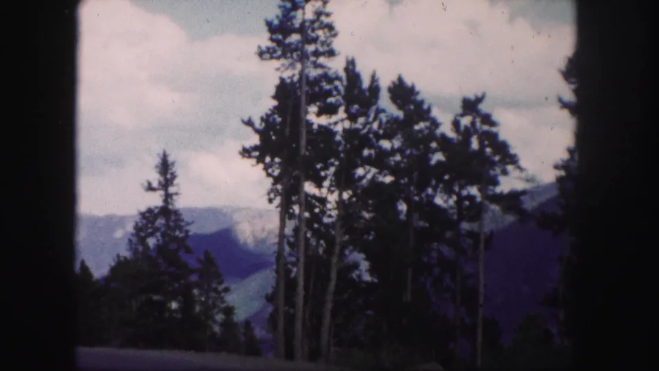 1958 Colorado 8mm frame showing tall evergreens in the foreground, layered mountain ridges, and a partly cloudy sky.