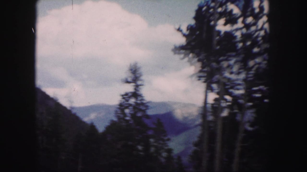 1958 Colorado 8mm frame showing dark evergreen trees in front of distant mountain ridges and a bright, cloudy sky.