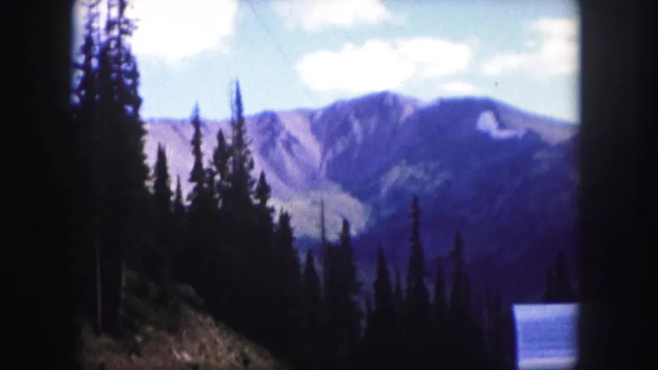 1958 Colorado 8mm frame showing rocky mountain ridges beyond dark evergreen silhouettes and a sloping forest foreground.