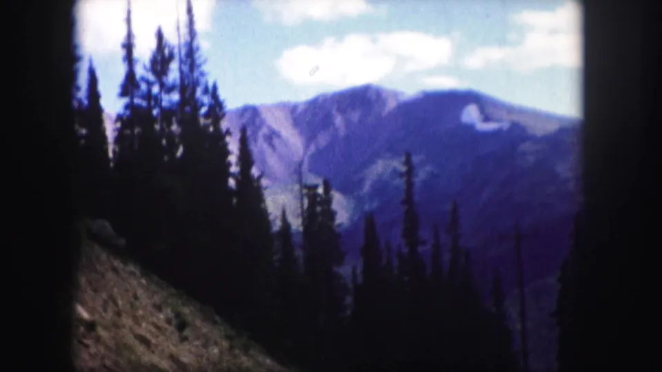 1958 Colorado 8mm frame showing evergreen trees along a hillside with distant mountain ridges under small white clouds.