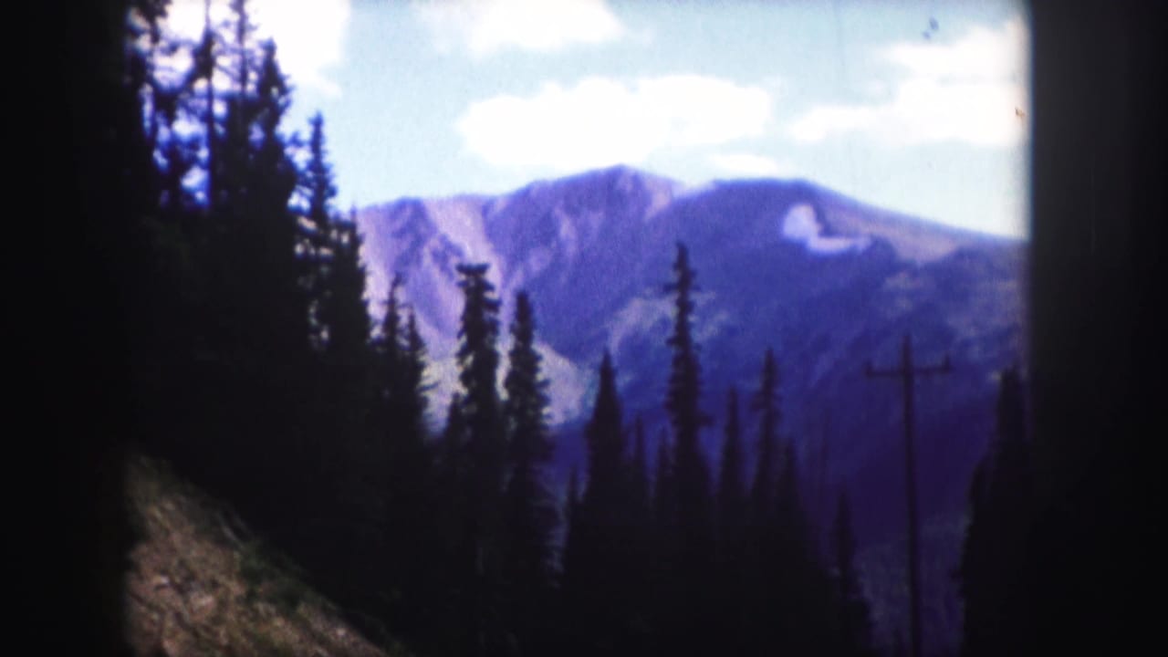 1958 8mm Colorado landscape view with dark evergreens in the foreground and blue-purple mountain ridges beyond.
