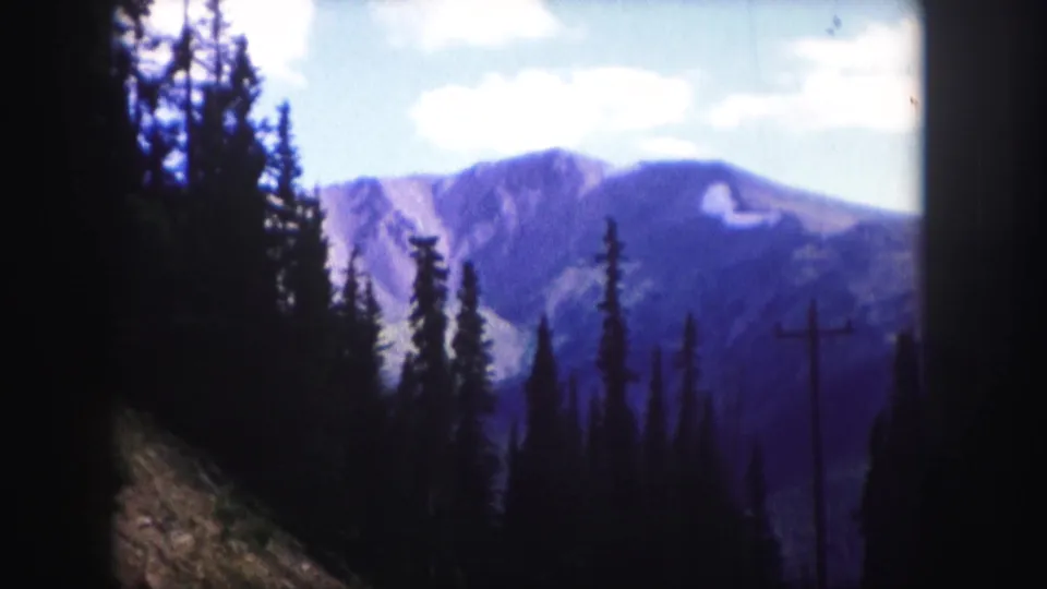 1958 8mm Colorado landscape view with dark evergreens in the foreground and blue-purple mountain ridges beyond.