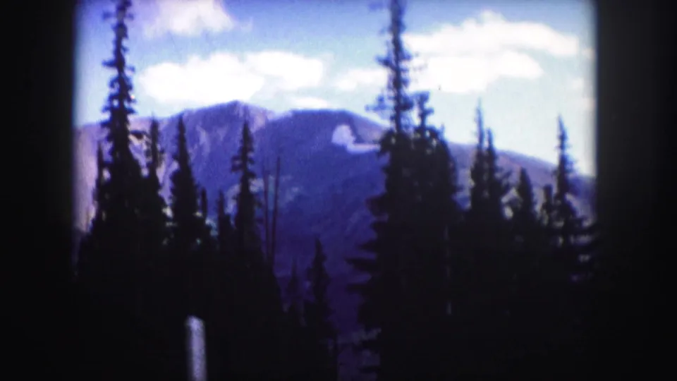 1958 Colorado home-movie frame with tall conifers silhouetted against a hazy blue mountain ridge and bright clouded sky.