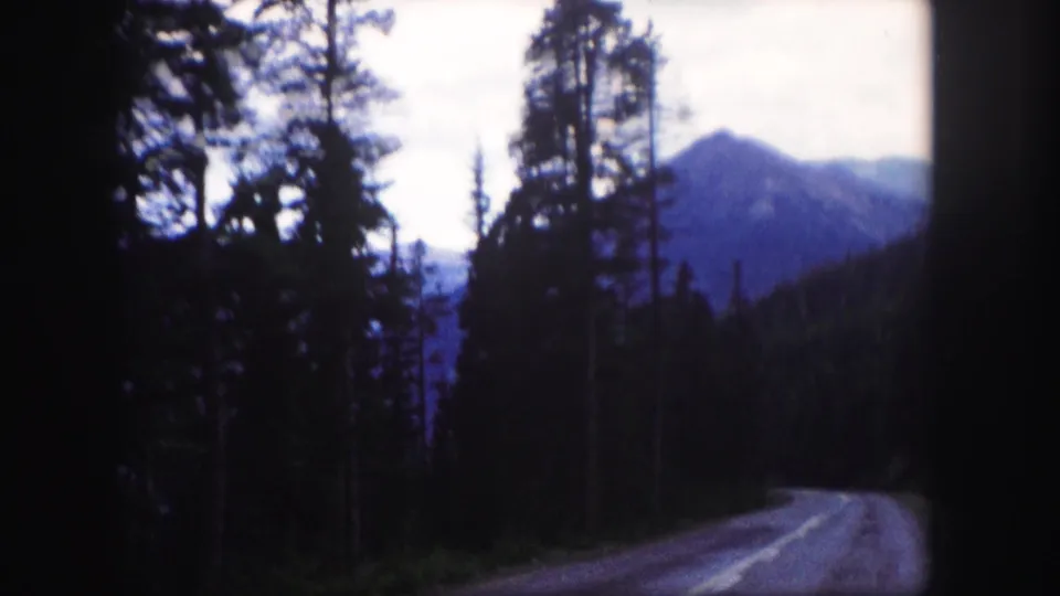 1958 Colorado 8mm frame with a paved road curving through forest toward a blue-toned mountain backdrop.