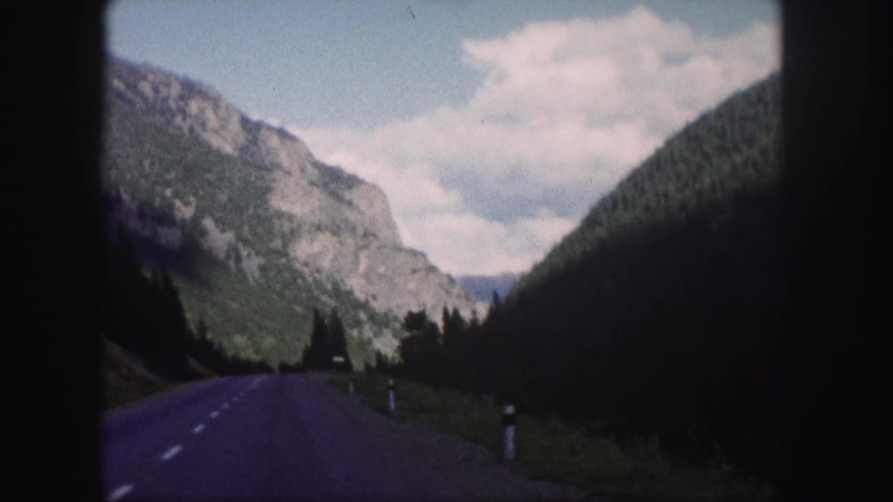 Mountain valley road bordered by forested slopes, rocky cliffs, roadside posts, and clouds.