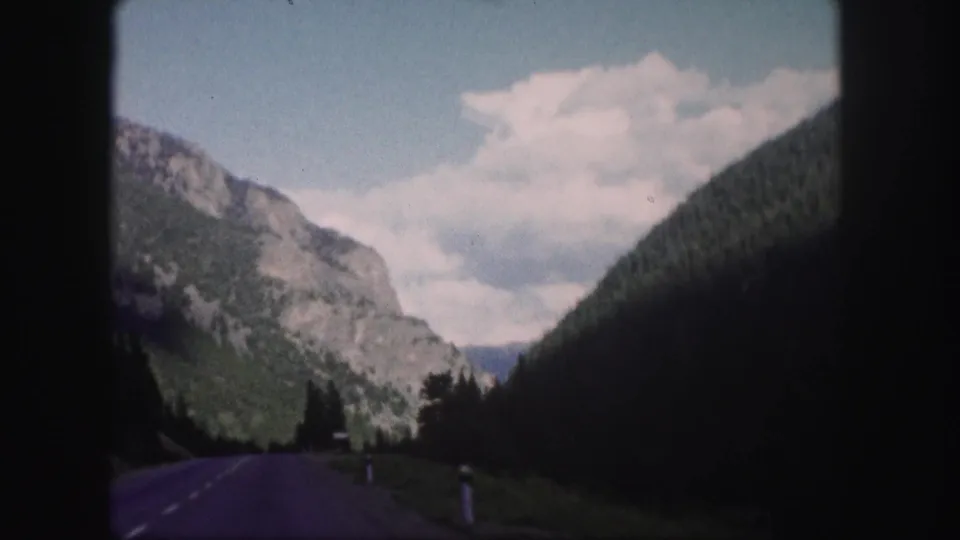 A moving-vehicle view of a Colorado mountain road, with a rocky cliff face on one side and dark wooded slope on the other.
