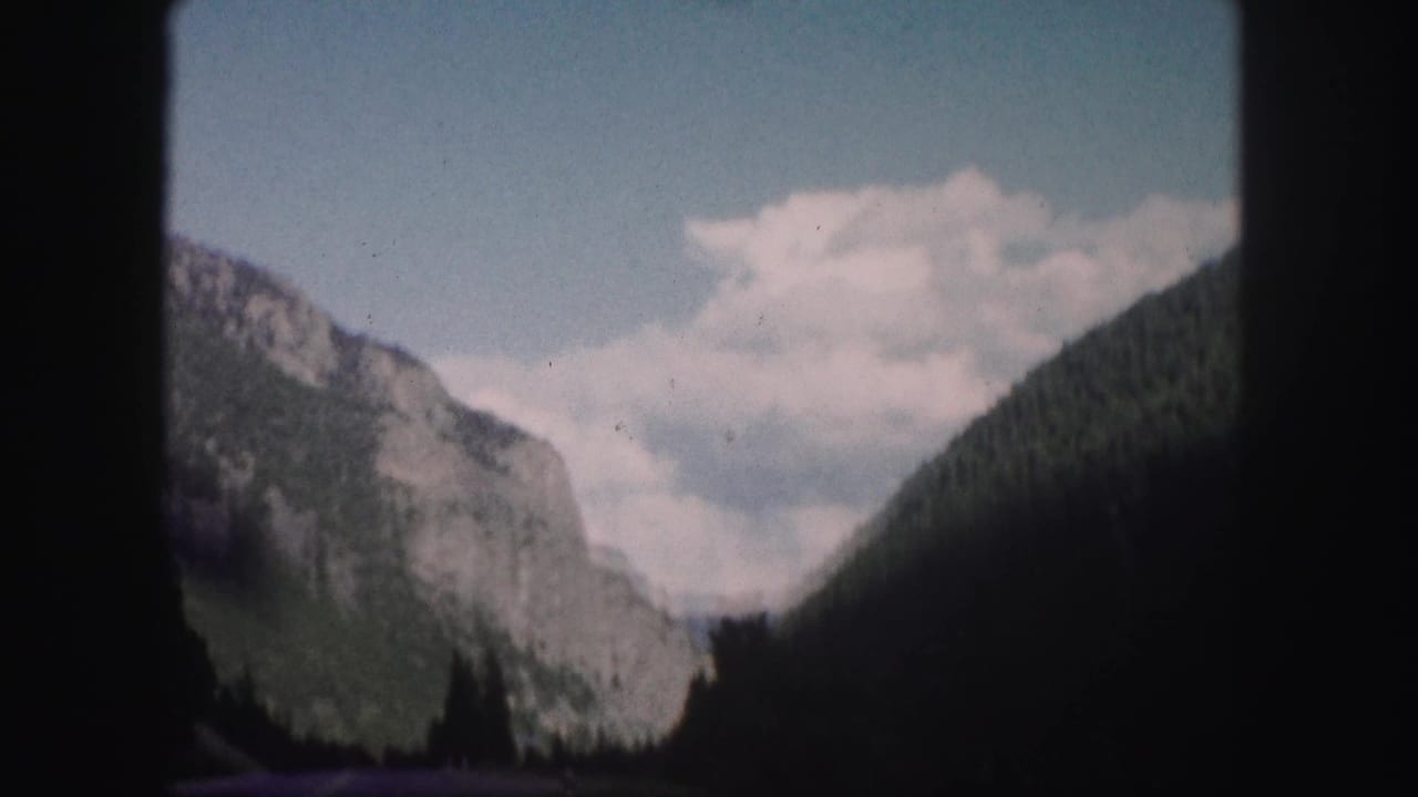1958 Colorado 8mm frame showing a mountain road approaching a rocky, forested valley beneath scattered clouds.