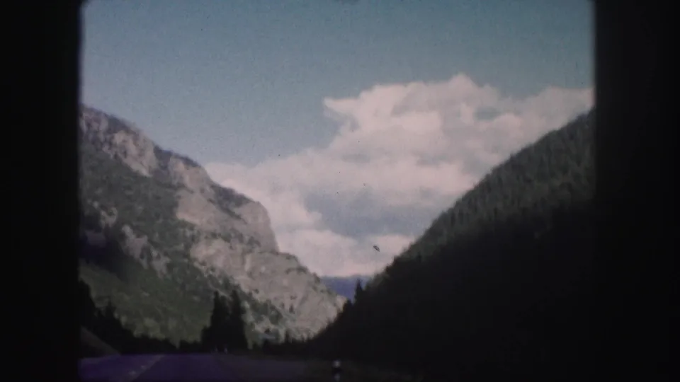 1958 Colorado road-travel frame with rocky slopes, dense trees, and a partly cloudy sky seen from a moving vehicle.