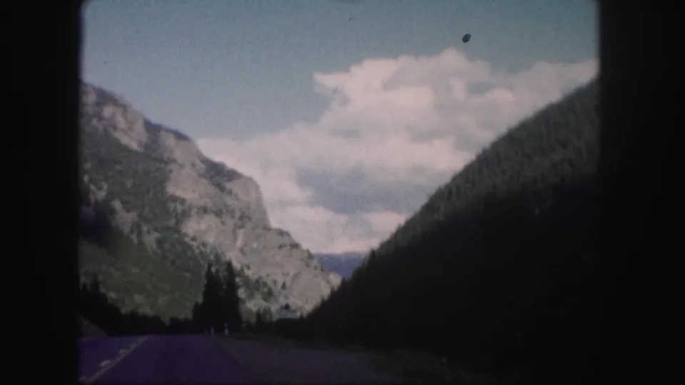 1958 Colorado 8mm view down a paved mountain road framed by steep tree-covered slopes and distant sky.