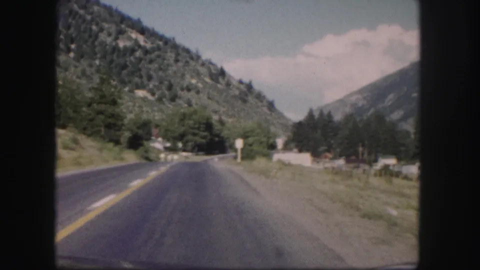 1958 Colorado driving view showing a paved highway, roadside posts, trees, and mountains rising on both sides.