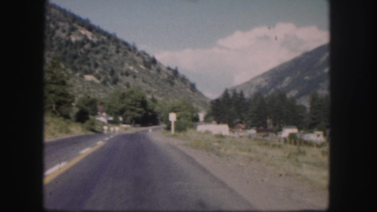 1958 Colorado 8mm frame showing a paved highway through a mountain valley with trees, signs, and scattered buildings.