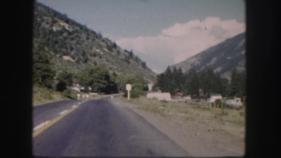 1958 Colorado 8mm frame showing a paved highway through a mountain valley with trees, signs, and scattered buildings.