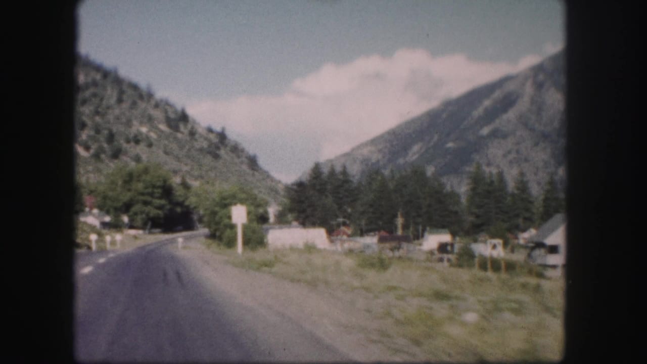 Mountain road beside open roadside ground, trees, signs, and small structures in a valley.