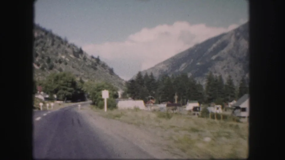 1958 Colorado road-trip frame looking along a two-lane highway past roadside signs and low buildings below forested mountains.