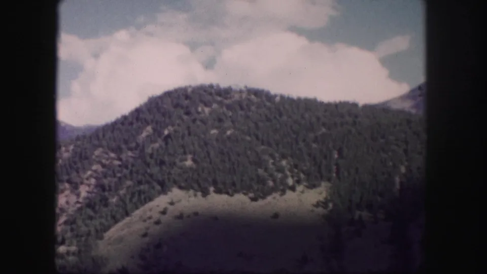 1958 Colorado 8mm frame of a forested mountain ridge and hillside beneath a bright clouded sky.