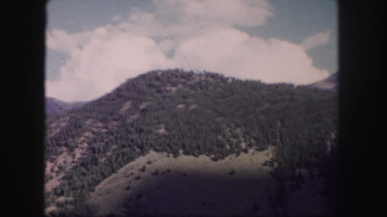 1958 8mm home-movie view of a forested Colorado mountain ridge beneath pale clouds and blue sky.