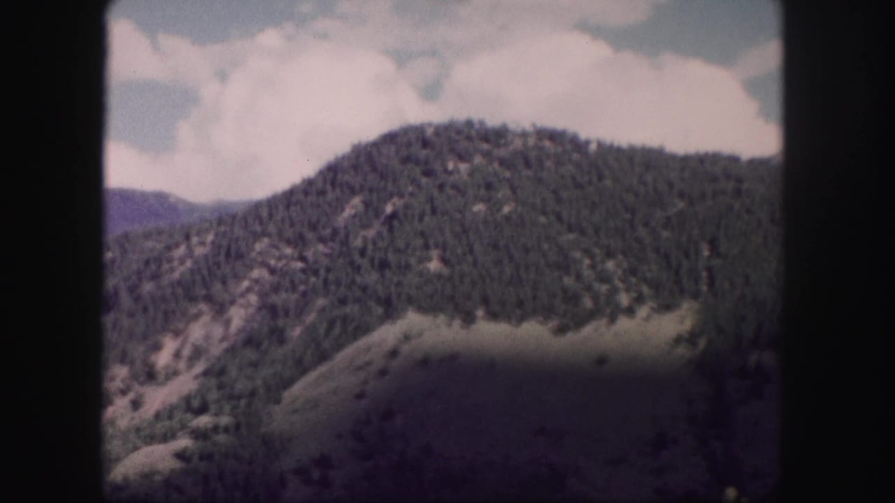 Rounded forested peak and shadowed lower slope beneath large white clouds.