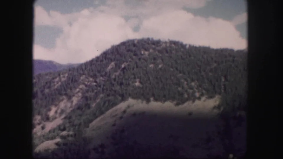 1958 Colorado mountain landscape with dense pine cover, a rounded summit, and soft home-movie color fading.