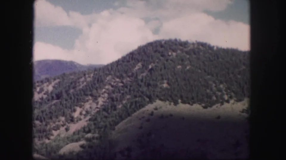 8mm frame of Colorado mountain slopes, with pine-covered terrain receding toward a distant ridge under cloudy sky.
