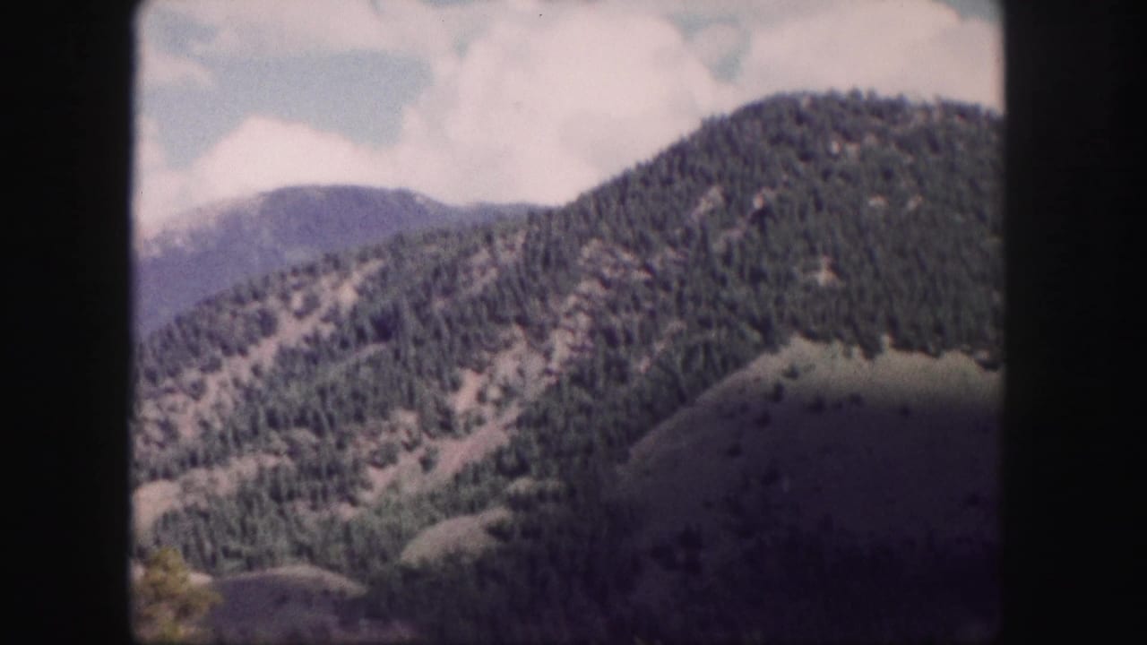 1958 Colorado 8mm frame showing forested mountain slopes and rounded hills under a bright sky.