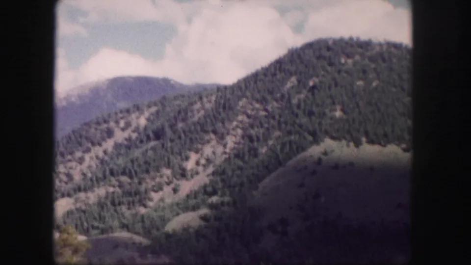 1958 Colorado 8mm frame showing forested mountain slopes and rounded hills under a bright sky.