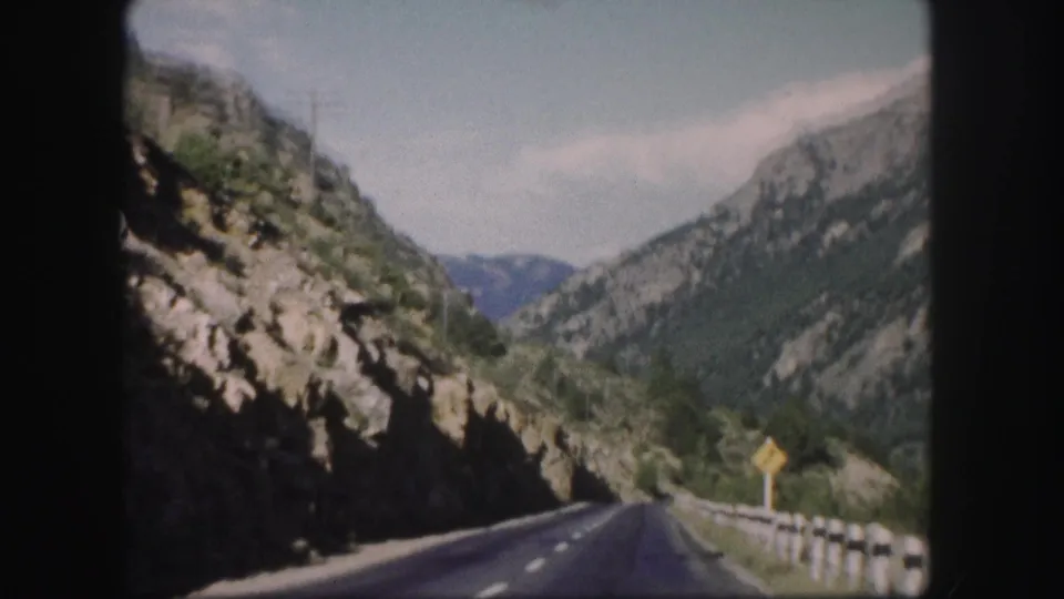 1958 Colorado 8mm road view with a guardrail, rocky canyon slope, evergreen hills, and a yellow roadside warning sign.