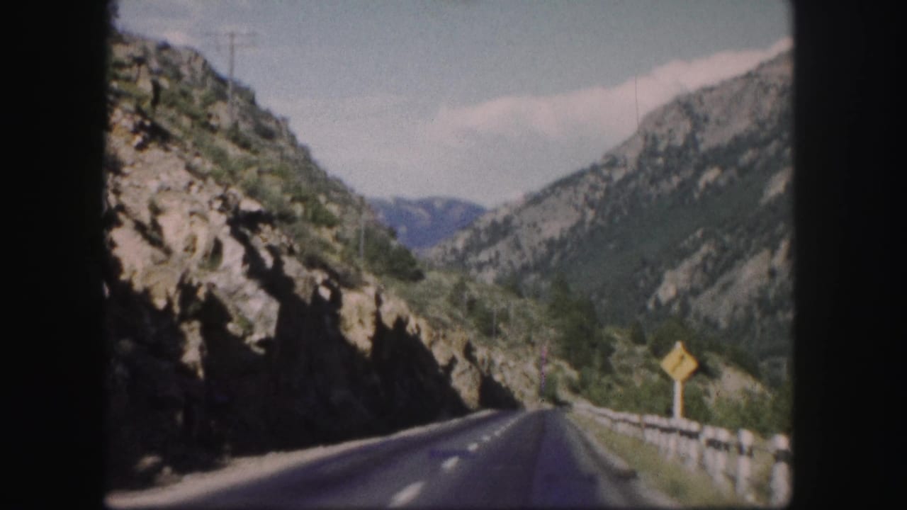 1958 Colorado 8mm view from a moving vehicle on a paved mountain road between rocky cliffs and forested slopes.