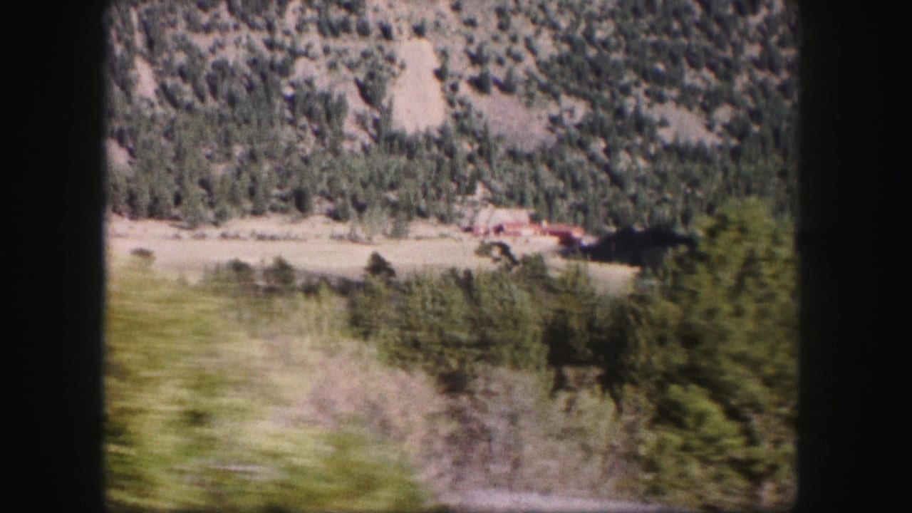 1958 8mm view of a green Colorado valley with trees, rocky slopes, and a small red-roofed building seen from a moving vehicle.