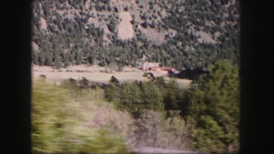 1958 8mm view of a green Colorado valley with trees, rocky slopes, and a small red-roofed building seen from a moving vehicle.