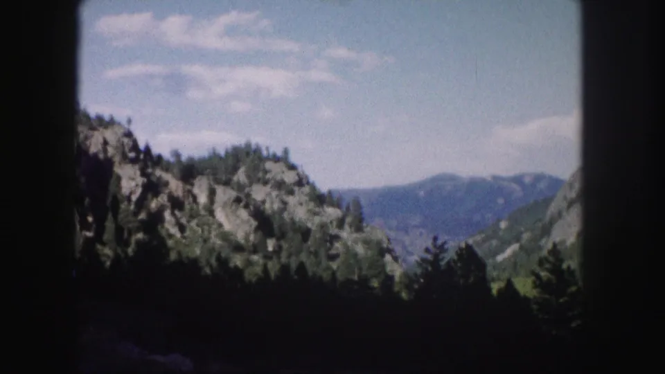 1958 Colorado 8mm frame shows a rocky forested slope, dark foreground trees, and distant blue mountains below pale clouds.