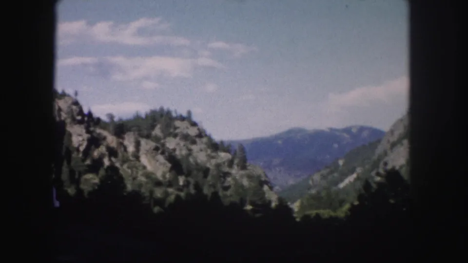 1958 Colorado home-movie frame with a distant mountain ridge, forested rocky slopes, pale sky, and window-edge shadow from a moving vehicle.