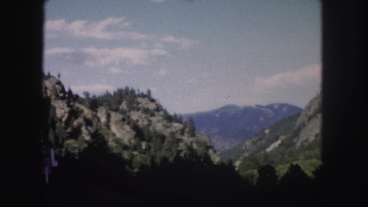 1958 Colorado home-movie frame showing forested rocky slopes, distant mountains, blue sky, and dark window edges from a moving car.