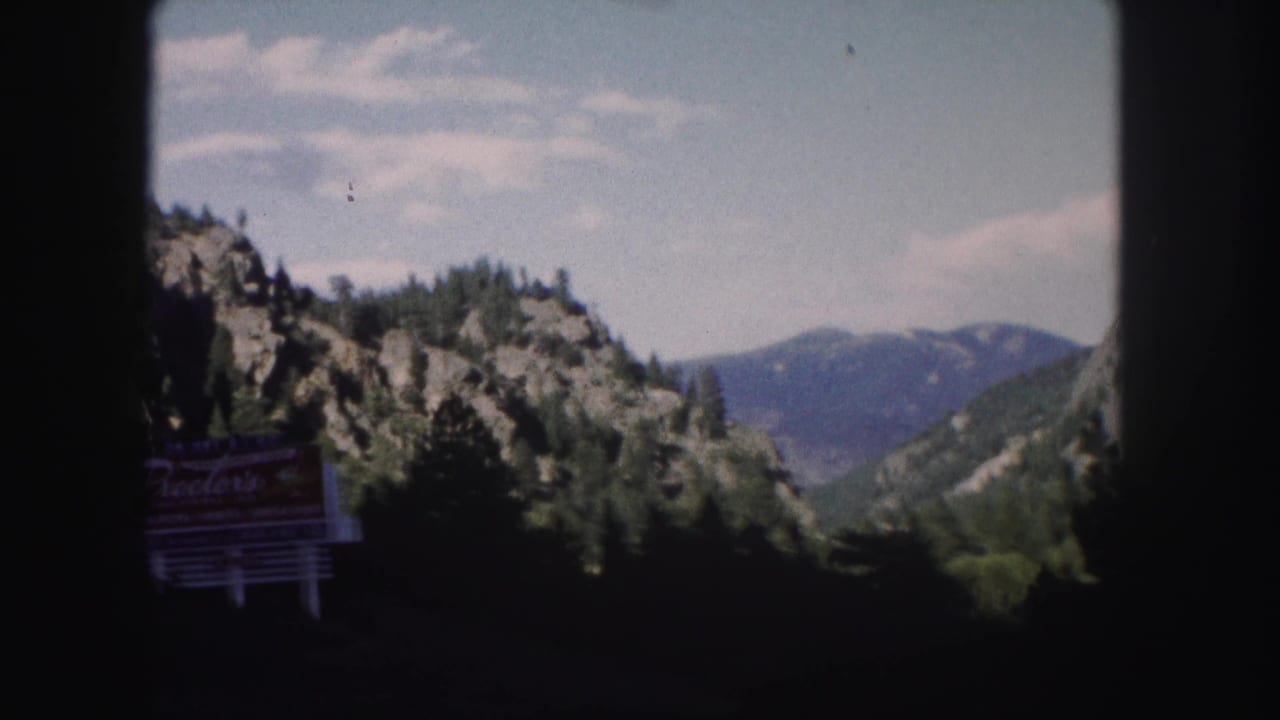 Roadside sign in shadow with rocky hills, trees, and mountains behind it.