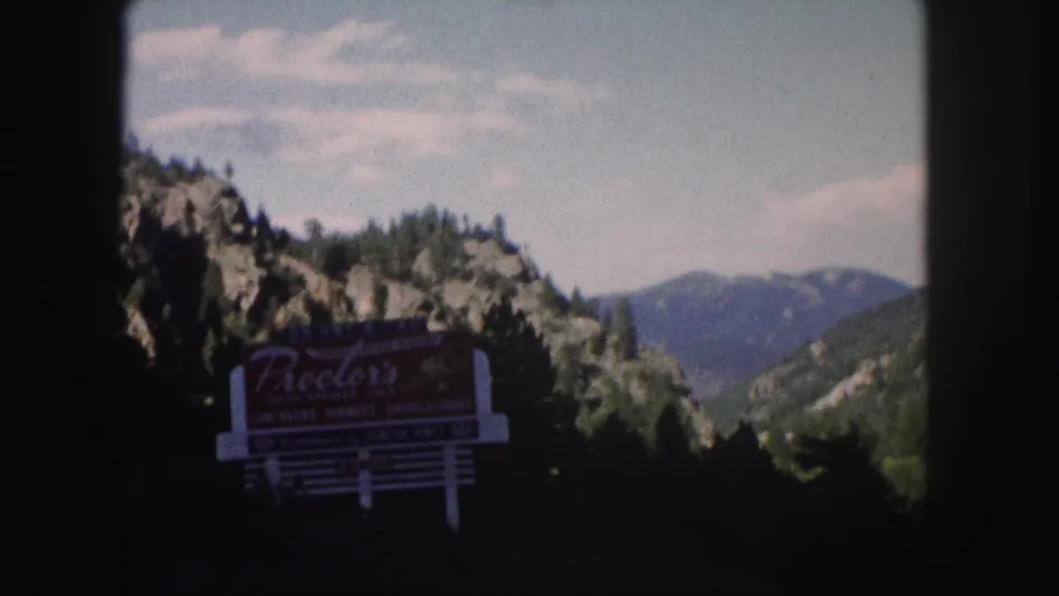 Colorado road-trip frame from 1958 showing a large roadside sign in shadow with wooded hills and mountains behind it.