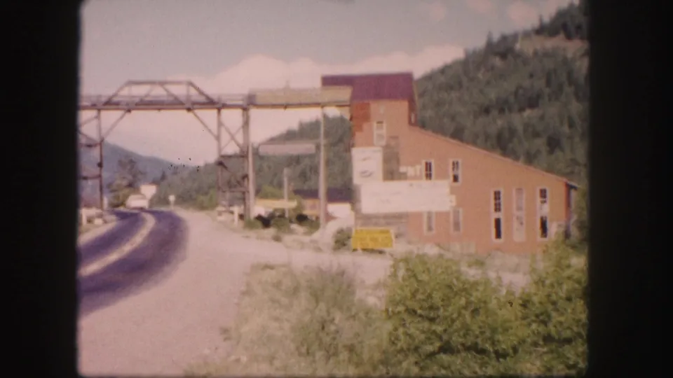 1958 Colorado roadside scene with a two-lane road, small building, overhead metal truss structure, brush, and wooded hills.