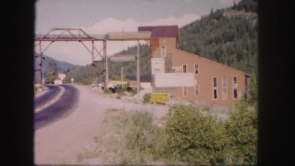 1958 Colorado 8mm frame showing a roadside industrial building, metal gantry, road shoulder, brush, and mountain greenery.