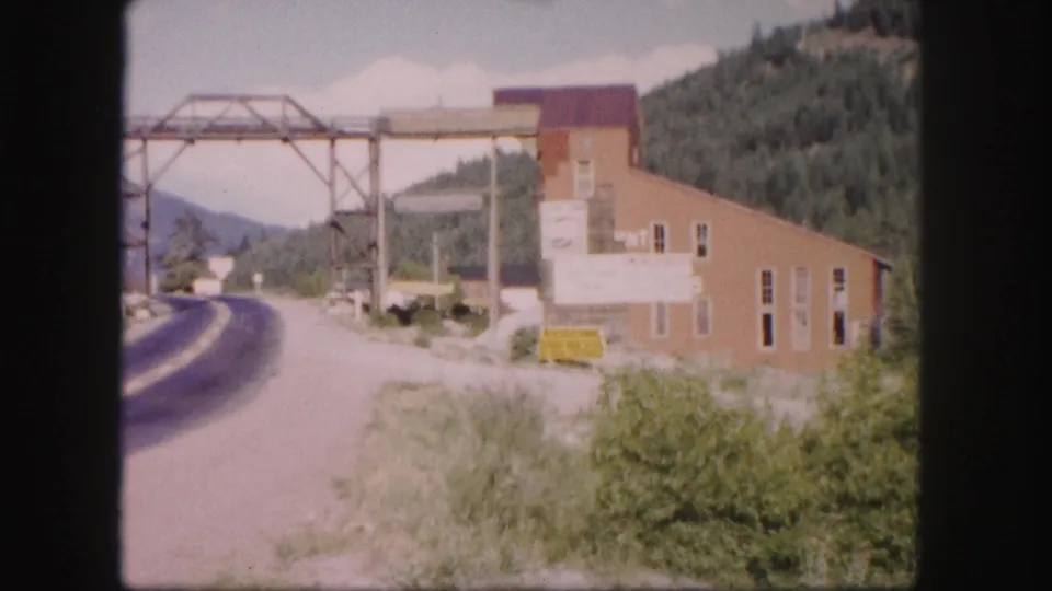 1958 Colorado frame with a curving roadway, brick industrial building, metal gantry, roadside vegetation, and wooded hills.