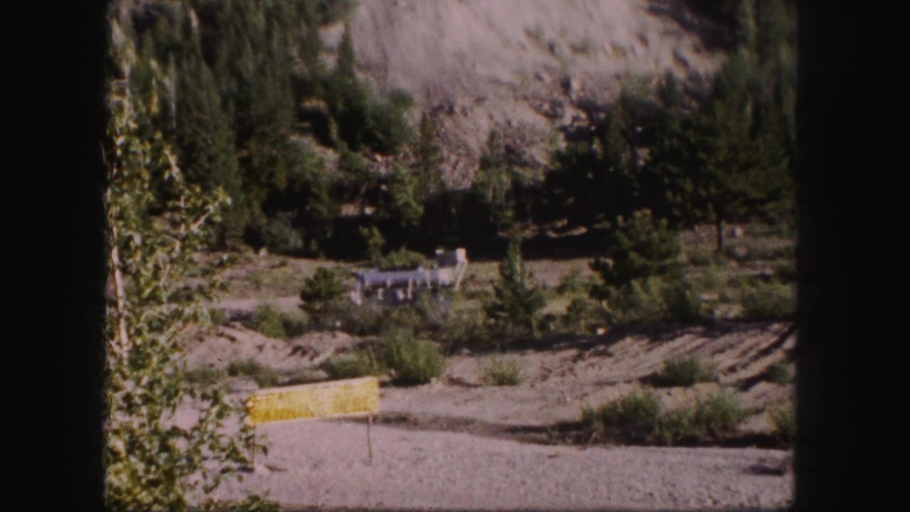 1958 Colorado 8mm frame showing a yellow roadside sign below evergreens, a rocky mountain slope, and a distant industrial structure.
