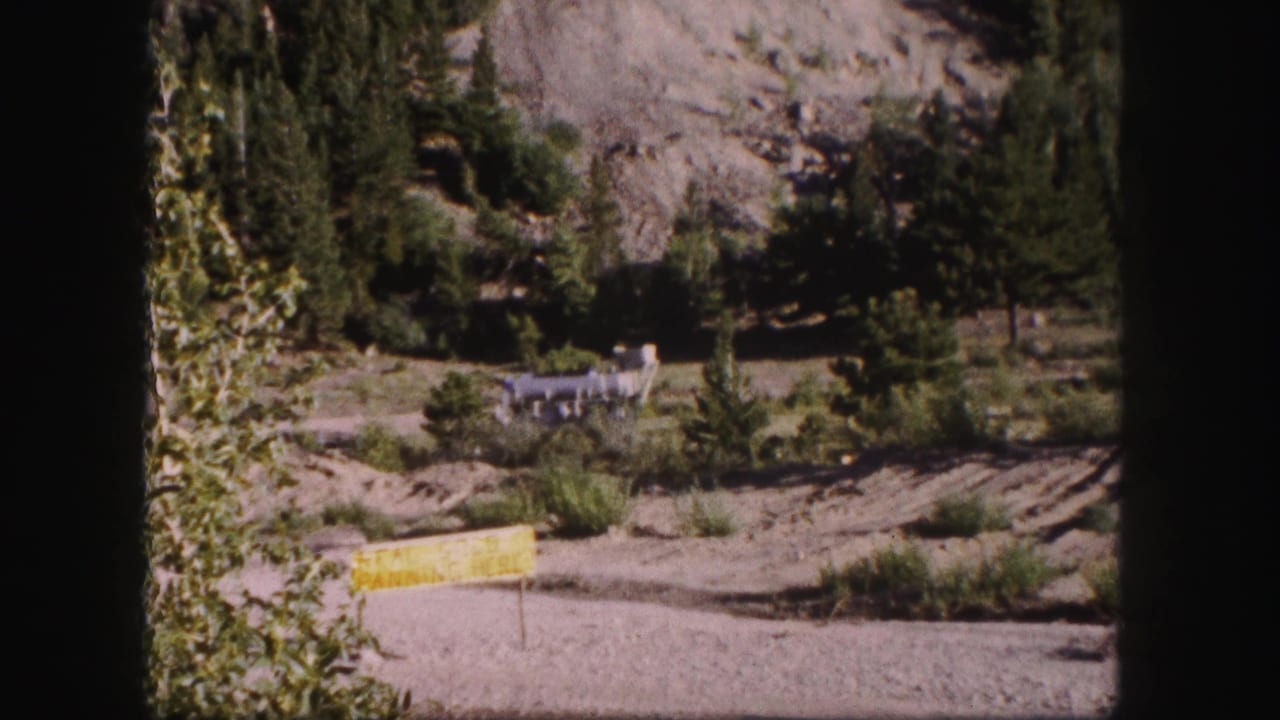 Evergreen mountain scene with a yellow foreground sign and distant industrial structure near a road.