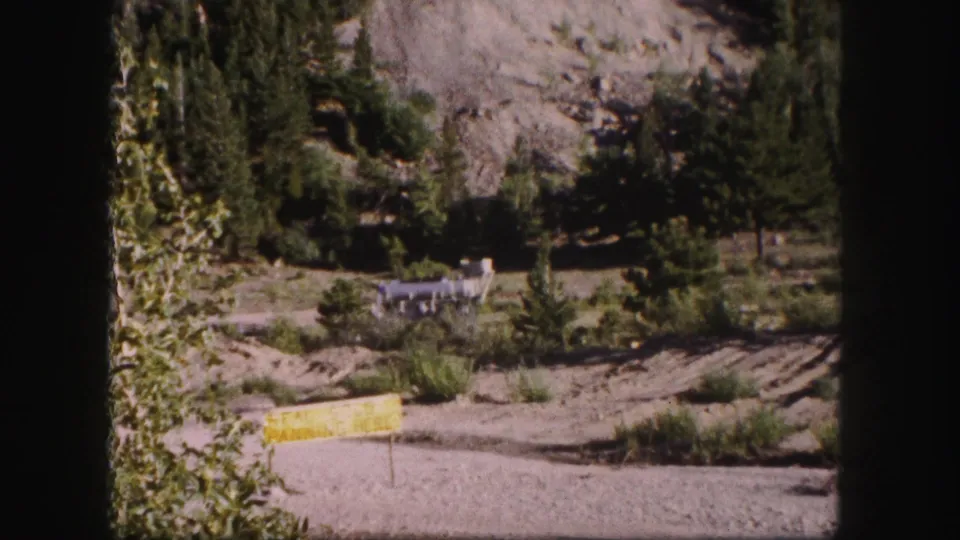 1958 Colorado mountain view with a yellow sign in the foreground, evergreens, rocky hillside, and distant industrial structure near the road.