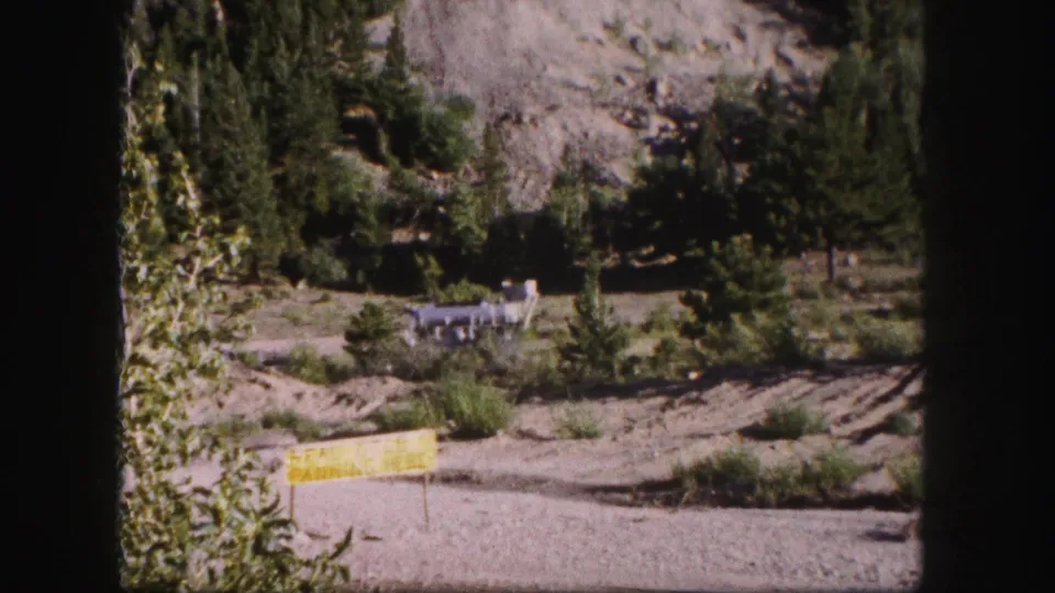 1958 Colorado 8mm frame with a yellow roadside sign, brush, evergreens, rocky slope, and distant industrial structure above the road.