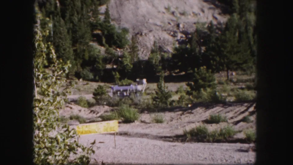 1958 Colorado home-movie frame showing a yellow sign in a gravel foreground with evergreens, rocky hillside, and distant industrial structure.