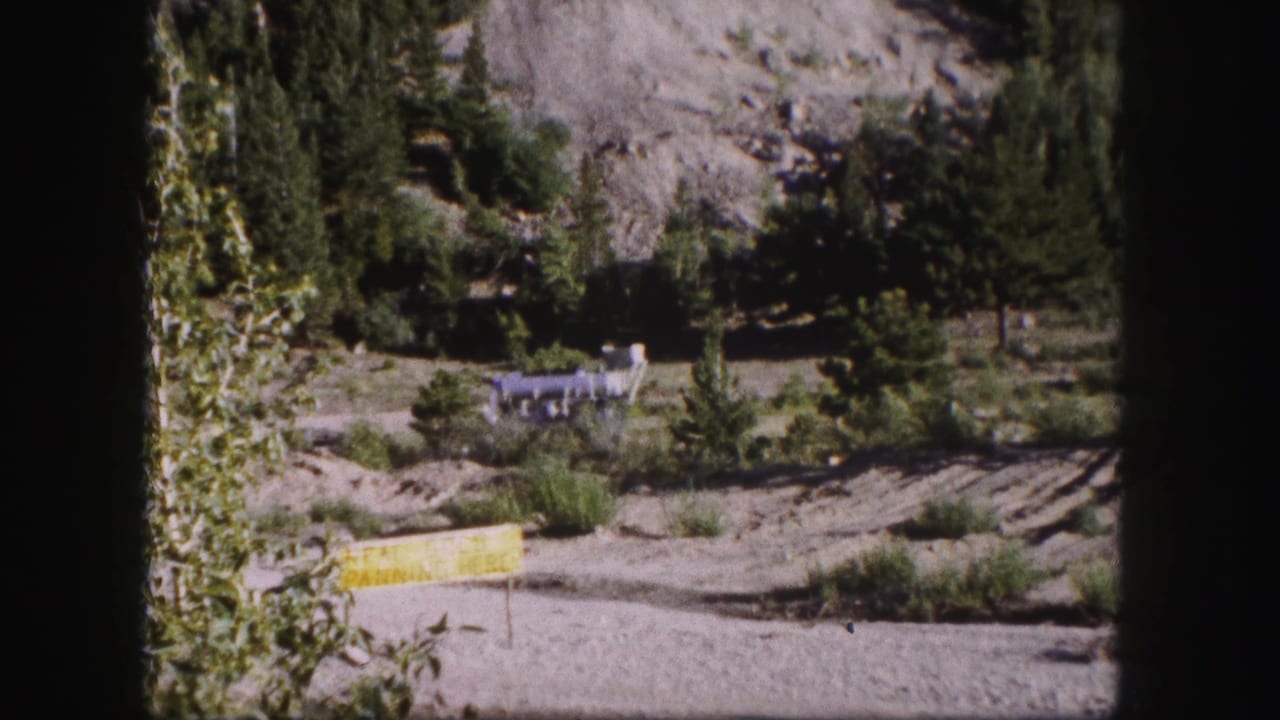 1958 Colorado 8mm frame showing a rocky mountain roadside, trees, a clearing, and a small distant structure.