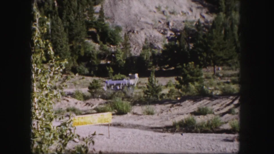 1958 Colorado 8mm frame showing a rocky mountain roadside, trees, a clearing, and a small distant structure.