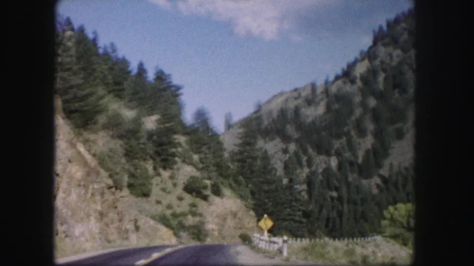 1958 Colorado 8mm frame of a mountain road curving past guardrail posts and a yellow roadside warning sign.