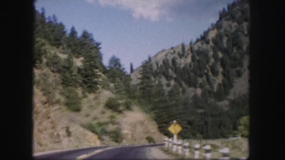 1958 Colorado home-movie frame showing a yellow curve sign, guardrail, and forested slopes along a mountain road.