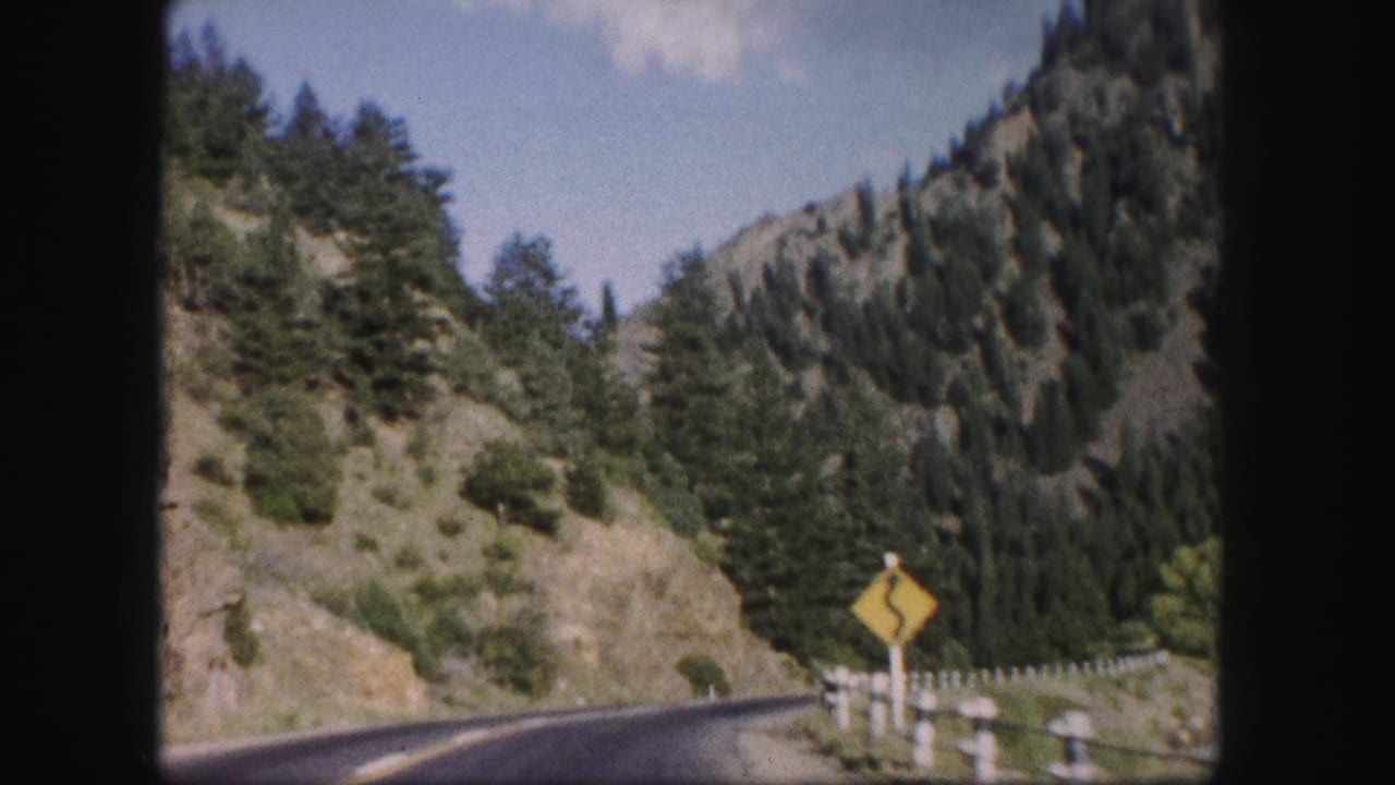 1958 Colorado 8mm frame of a winding mountain road passing a yellow curve sign, guardrail, rocky hillside, and dense evergreens.